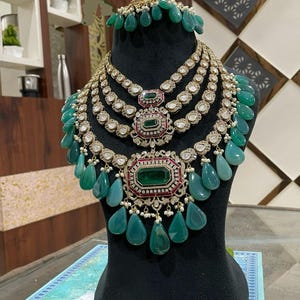 May include: A multi-layered emerald green and diamond necklace with a large emerald green pendant. The necklace is on a black display stand.