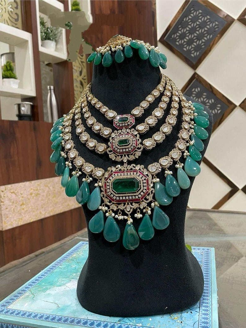 May include: A multi-layered emerald green and diamond necklace with a large emerald green pendant. The necklace is on a black display stand.