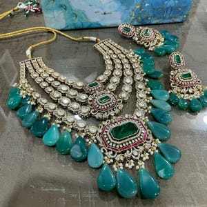 May include: A multi-layered gold necklace with emerald green and pink stones. The necklace has a teardrop design with multiple strands of pearls and diamonds. The necklace is paired with matching earrings.