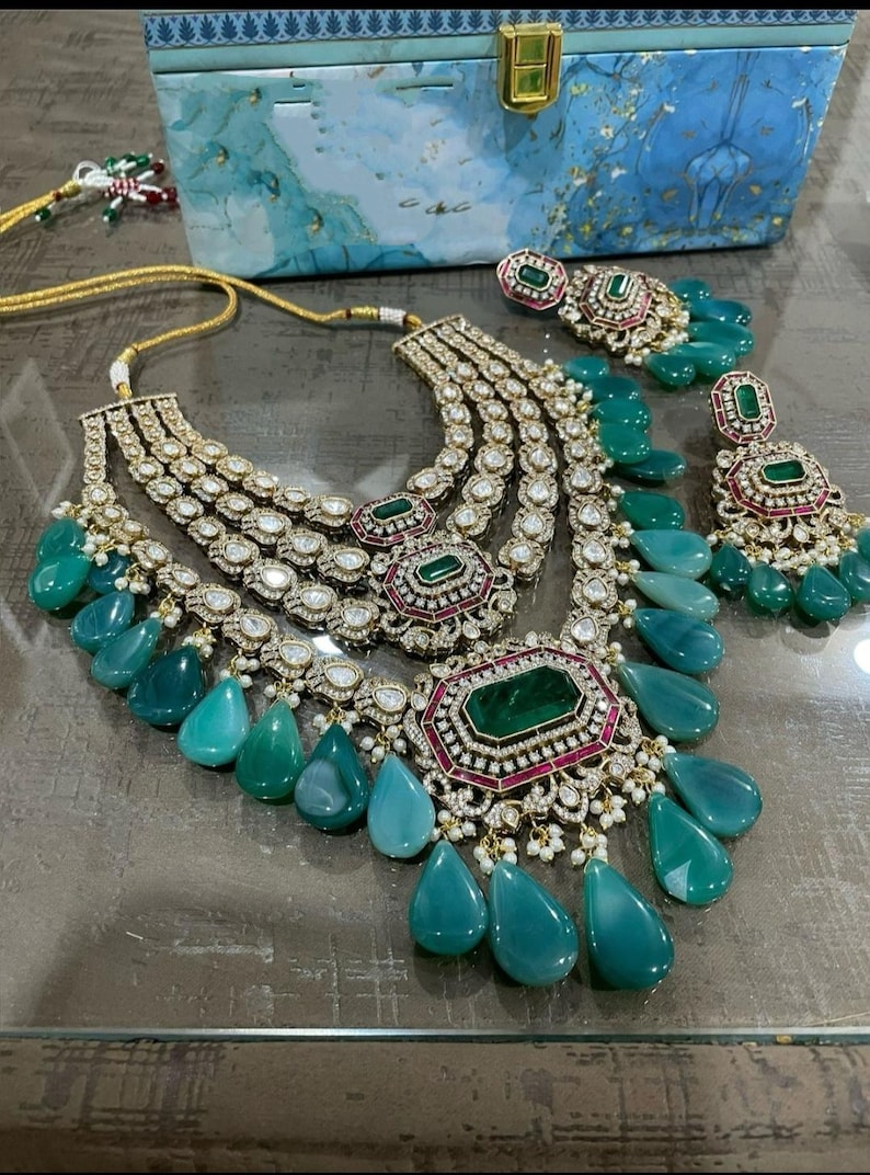 May include: A multi-layered gold necklace with emerald green and pink stones. The necklace has a teardrop design with multiple strands of pearls and diamonds. The necklace is paired with matching earrings.