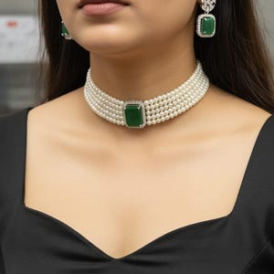 May include: A pearl choker necklace with a rectangular emerald green stone at the center, surrounded by small clear stones. Matching emerald green and clear stone earrings are also visible. The jewelry is set against a black top.