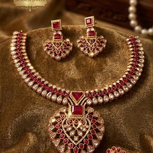 May include: A gold-toned jewelry set featuring a necklace, earrings, and a ring. The necklace has a heart-shaped pendant with red and white gemstones. The earrings and ring also feature red gemstones. The set is displayed on a brown surface.