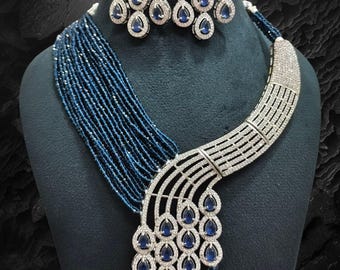 May include: A dazzling jewelry set featuring a necklace and matching earrings. The necklace has a silver-toned, curved design with clear stones and dangling blue teardrop-shaped gems. The earrings mirror the design. The necklace also has strands of blue beads.