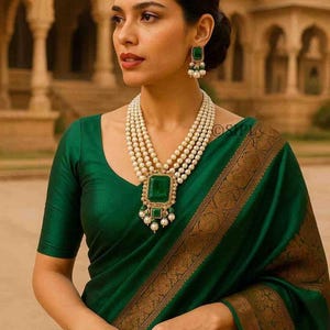 Può includere: Una donna indossa un sari verde scuro con bordo dorato. Indossa una collana di perle a più fili con un grande pendente di smeraldo e orecchini abbinati. Il sari e i gioielli creano un look elegante e formale.