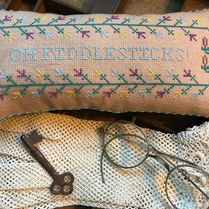 May include: A beige pillow with a cross-stitch design featuring the text "Oh Fiddlesticks!" surrounded by a green vine border. The pillow is sitting on a white crocheted doily with a vintage key and round glasses.