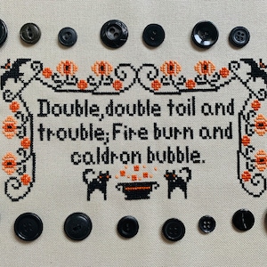 May include: A black and white cross-stitch embroidery with the text "Double, double toil and trouble; Fire burn and caldron bubble." surrounded by black buttons.
