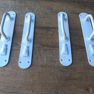 Könnte beinhalten: Vier weiße Türgriffe mit geschwungenem Griffdesign. Jeder Griff ist auf einer rechteckigen Grundplatte mit einem dekorativen Element an der Basis montiert. Die Griffe sind auf einer Holzoberfläche angeordnet.