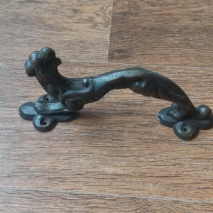 Puede incluir: Un tirador de puerta de color bronce oscuro con un diseño curvo y ornamentado. El tirador presenta una figura decorativa con forma de animal en la parte superior y está montado sobre dos placas decorativas con orificios para tornillos. Para puerta.