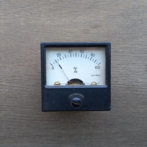 Könnte beinhalten: Ein schwarzes analoges Messgerät mit weißem Zifferblatt und einer Nadel, die einen Wert nahe Null anzeigt. Das Zifferblatt zeigt Prozentsätze von 0 bis 100 an, mit dem Prozentzeichen. Das Messgerät hat einen kleinen Einstellknopf.