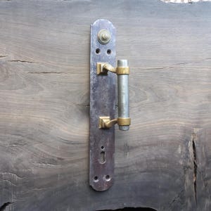 May include: A vintage door handle with a distressed metal finish. The handle features a long, rectangular backplate with a cylindrical handle and square accents in brass. The handle is mounted on a weathered wooden door.