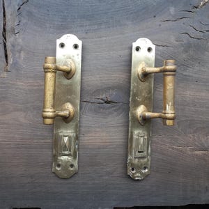 May include: Two antique brass door handles. Each handle has a long, rectangular backplate with a decorative handle and a small, rectangular detail at the bottom. The handles are a warm, aged brass color, showing signs of wear.