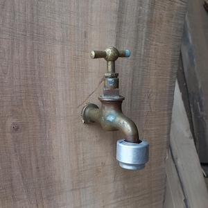 May include: A vintage brass faucet with a cross-shaped handle is mounted on a weathered wooden surface. The faucet has a curved spout and a silver-colored cap at the end. The wood has a natural grain.