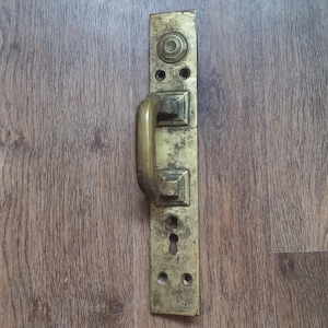 May include: A vintage brass door handle with a rectangular backplate. The handle is a curved bar, and the backplate features a keyhole and decorative details. The brass has a weathered appearance, suggesting age and use. The door hardware is likely from an older home.