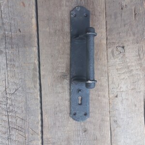May include: Black metal door handle with a keyhole and a long, cylindrical handle. The handle is mounted on a wooden surface.