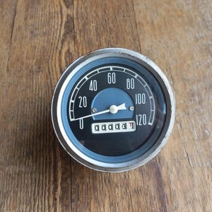 Könnte beinhalten: Ein Vintage-Tachometer mit schwarzem Zifferblatt, weißen Ziffern und einem blauen Zentrum. Das Messgerät zeigt bis zu 120 mph (ca. 193 km/h) an. Der Kilometerzähler zeigt 000031 an. Das Metallgehäuse hat eine silberne Oberfläche. Der Hintergrund ist eine Holzoberfläche.