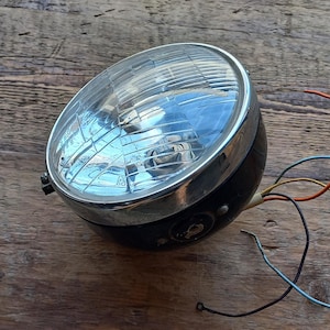 May include: A vintage motorcycle headlight with a chrome trim and a black housing. The headlight has a clear glass lens with a ribbed pattern. The headlight is connected to a set of wires.