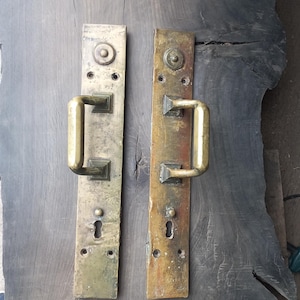 Może przedstawiać: Dwa antyczne mosiężne uchwyty do drzwi z otworami na klucze i prostokątnymi płytkami tylnymi. Uchwyty mają zakrzywioną, prostokątną konstrukcję. Mosiądz ma zwietrzały, postarzany wygląd, z różnymi odcieniami. Zamontowane na ciemnej drewnianej powierzchni.