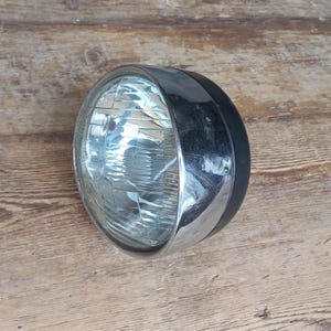 May include: A chrome motorcycle headlight with a black rubber housing. The headlight has a clear glass lens with a grid pattern.
