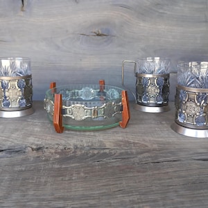 May include: Set of four glass teacups with metal holders and a glass bowl with a wooden base. The teacups and bowl have intricate designs.