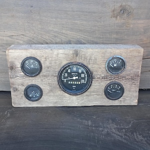 May include: A rustic wooden plaque with five vintage car gauges. The gauges are black with white markings and needles. The gauges measure speed in kilometres per hour, fuel level, oil pressure, temperature, and battery voltage.