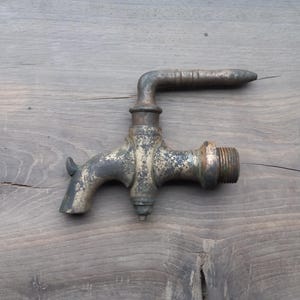 Può includere: Un antico rubinetto in ottone dall'aspetto usurato. Il rubinetto ha un beccuccio curvo, un'impugnatura e un'estremità filettata per il fissaggio. Il rubinetto è posto su uno sfondo di legno.