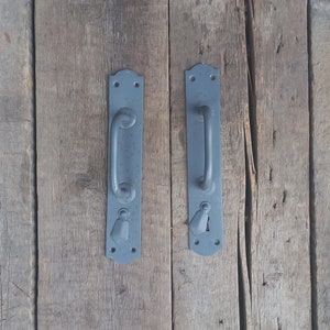 Może przedstawiać: Dwie szare metalowe klamki do drzwi z ozdobnym wzorem. Klamki są zamontowane na drewnianej powierzchni.