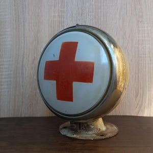 Könnte beinhalten: Ein kugelförmiges medizinisches Schild mit einer weißen Vorderseite und einem markanten roten Kreuzsymbol. Das Objekt hat einen metallischen, verwitterten Rahmen und ist auf einem runden Sockel montiert, was ihm ein altes Aussehen verleiht.