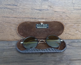 May include: A pair of round sunglasses with black frames and tinted lenses are displayed in an open brown leather case. The case has a soft interior. The text "Rudolf Jacob Uhrmacher & Optiker Netzschkau" is printed on the case.