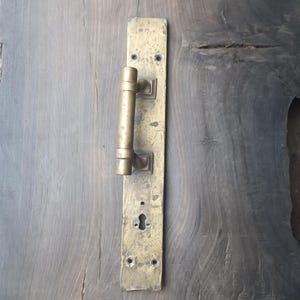 May include: A weathered brass door handle and plate are mounted on a gray wooden door. The handle is cylindrical, and the plate has a keyhole and screw holes. The wood grain is visible.