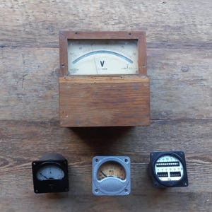 Puede incluir: Tres voltímetros analógicos vintage con caras negras y plateadas. El voltímetro superior está en una caja de madera y tiene una aguja que apunta a 90. Los otros dos voltímetros tienen cajas negras y plateadas y están etiquetados como "A" y "MΩ".