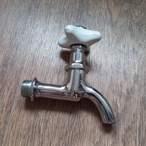 May include: A chrome-plated metal water faucet featuring a star-shaped white handle at the top. The faucet has a threaded inlet pipe on the left side and a curved spout pointing downward on the right side, with a smooth, reflective finish.