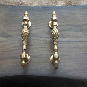 May include: Two gold-toned door handles with a decorative, ornate design. The handles have a pointed top and a round base.