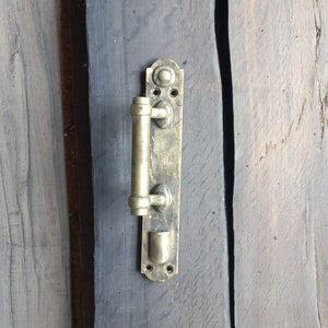 May include: Close-up of an antique brass door handle mounted on a weathered, dark gray wooden door. The handle has a cylindrical grip and a rectangular backplate with decorative details. The wood grain is visible.
