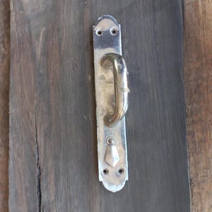 May include: A vintage door handle with a silver-colored backplate and a curved, brass-colored handle. The handle is mounted on a dark wooden door. The backplate has two screw holes at the top and bottom, and a decorative diamond shape near the bottom.