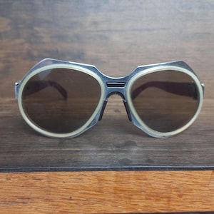 May include: Sunglasses featuring a metallic silver frame with a distinct angular top edge and rounded dark lenses. Each lens is encircled by a thick, cream-colored border that provides a contrasting outline against the metallic structure of the eyewear.