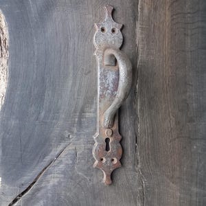 May include: A weathered, silver-toned door handle with an ornate design and a keyhole. The handle is attached to a dark wooden door.