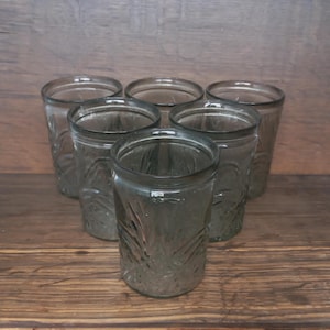 Puede incluir: Conjunto de seis vasos para beber transparentes teñidos de gris. Cada vaso tiene una forma cilíndrica con un patrón en relieve texturizado. Los vasos están vacíos y parecen ser de vidrio. El fondo es una superficie de madera.