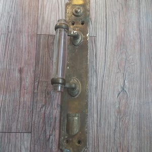 May include: A vintage door handle assembly featuring a long, rectangular brass backplate with a weathered patina, two mounting holes, and a clear, cylindrical glass grip held in place by two brass supports and a small lower brass cylinder.
