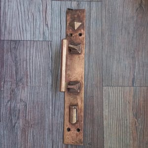 Può includere: Una piastra rettangolare per maniglia in ottone con una barra di trazione verticale, un accento piramidale superiore, due supporti di montaggio quadrati e una fessura per chiave inferiore. La superficie metallica presenta una patina invecchiata.