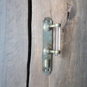 Może przedstawiać: Uchwyt do drzwi w stylu vintage z prostokątną mosiężną płytką tylną i przezroczystym akrylowym uchwytem. Uchwyt jest zamontowany na zwietrzałych drewnianych drzwiach, prezentując naturalne usłojenie i fakturę drewna. Mosiądz ma patynę.