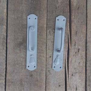 May include: Two gray metal door handles with a simple design. The handles have a rounded top and bottom and a curved handle in the center. The handles are mounted on a wooden surface.