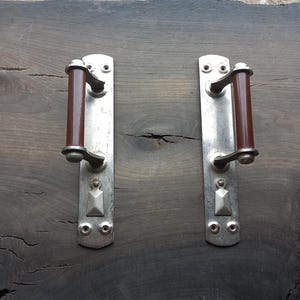 Könnte beinhalten: Zwei antike Türgriffe mit silberfarbenen Metallplatten und braunen zylindrischen Griffen. Jede Platte hat eine Schlüssellochform und dekorative Nieten. Die Griffe sind auf einer dunklen Holzoberfläche angebracht.