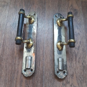 May include: Two matching door handles featuring a long, rectangular polished metal backplate with rounded ends. Each handle has a cylindrical black grip supported by two brass-toned metal brackets and a small, silver-toned cylindrical latch mechanism.