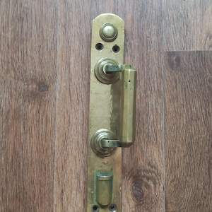 Puede incluir: Un tirador y placa de puerta de latón antiguo, montados en una puerta de madera. El tirador es rectangular con un agarre redondeado. La placa tiene detalles decorativos y orificios de montaje. El latón tiene una pátina cálida y envejecida.