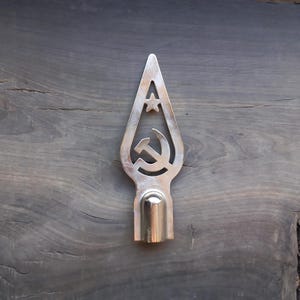 May include: A silver-colored metal spearhead with a cut-out design featuring a star and a hammer and sickle symbol. The spearhead has a rounded base and is set against a dark wood grain background.
