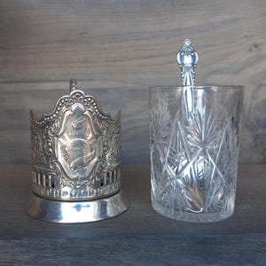 Puede incluir: Un portavasos de té plateado con detalles ornamentados y un vaso transparente con una cuchara. El vaso tiene un patrón de estrella. El soporte tiene un escudo con texto y un diseño de globo.