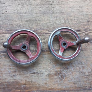 Könnte beinhalten: Zwei Vintage-Metallhandräder mit rot lackierten Speichen und silbernen Felgen. Jedes Rad hat einen silbernen Griff. Die Farbe weist Gebrauchsspuren auf, was ihnen ein rustikales Aussehen verleiht. Diese Räder stammen wahrscheinlich von Maschinen oder Geräten.
