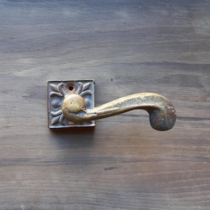 Könnte beinhalten: Antiker Türgriff aus Messing mit quadratischer Grundplatte mit floralen Details. Der Griff hat ein gebogenes Design und weist Alters- und Gebrauchsspuren mit Patina-Finish auf. Die Hardware ist auf einer Holzoberfläche montiert.