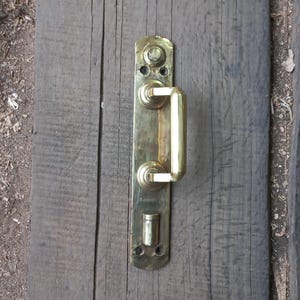 Op de afbeelding: Een messing deurklink met een rechthoekige achterplaat en een gebogen handvat. De klink is gemonteerd op een verweerd houten oppervlak, wat het vintage ontwerp benadrukt. Het messing heeft een gepolijste afwerking.