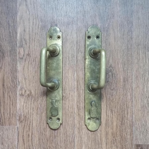 Op de afbeelding: Twee antieke messing deurklinken. Elke klink heeft een verticale rechthoekige achterplaat en een gebogen handvat. De klinken zijn gemonteerd op een houten oppervlak met een houtnerfpatroon. Het messing heeft een licht verouderde uitstraling.